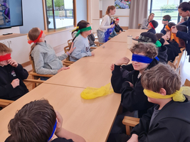 In einem hellen Raum sitzen Kinder an einem großen Tisch. Einige tragen bunte Tücher über den Augen, während Erwachsene ihnen Anweisungen geben oder sie beobachten. An einer Wand ist ein Bildschirm sichtbar und es gibt eine freundliche, spielerische Atmosphäre.