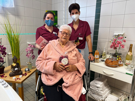 Eine Seniorin in einer pinken Decke, umgeben von Pflegekräften in einem gemütlichen Raum.