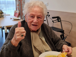 Eine ältere Frau mit einem Daumen hoch sitzt am Tisch mit einer Schüssel Suppe.