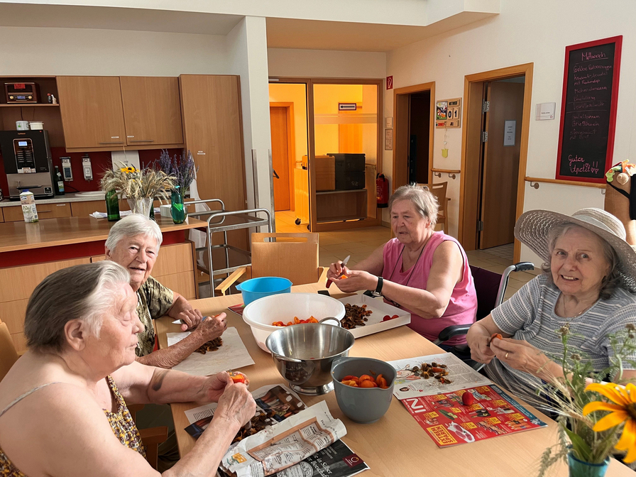 Vier ältere Frauen sitzen an einem Tisch in einem hellen Raum und bereiten Obst vor. Sie schälen oder schneiden Früchte und verwenden Schalen und eine große Schüssel. Der Tisch ist mit bunten Zeitschriften und einer Vase mit Blumen dekoriert.