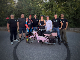 Eine Gruppe von acht Personen steht um ein pinkfarbenes Moped. Im Hintergrund sind Bäume zu sehen. Alle tragen verschiedene Kleidung, einige haben Melodien- oder Motorradkleidung an. Auf dem Boden ist ein Reifenabdruck zu sehen.