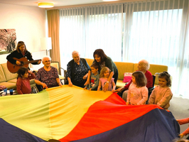 Eine Gruppe von älteren Frauen und Kindern sitzt im Raum und hält ein großes, farbenfrohes Tuch. Eine Frau spielt Gitarre, während die anderen mit dem Tuch spielen. Der Raum ist hell und freundlich gestaltet.
