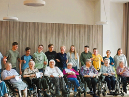 Junge Menschen stehen lächelnd hinter Seniorinnen in Rollstühlen.