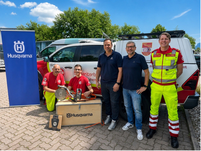 Fünf Personen stehen in einem Außengelände neben einem Fahrzeug. Zwei Personen tragen rote Shirts, während die anderen in Zivilkleidung bzw. Arbeitskleidung gekleidet sind. Vor ihnen befindet sich eine Kiste mit Werkzeugen und eine Fahne mit dem Husqvarna-Logo im Hintergrund.