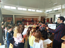 Kinder singen für Senioren in einem Gemeinschaftsraum. Ein Musiker begleitet sie.