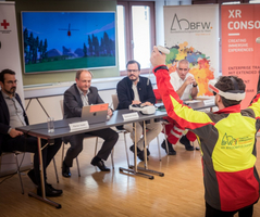 Ein Mann in Warnkleidung demonstriert Virtual-Reality-Technologie vor einer Gruppe am Tisch.