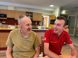 Ein älterer Mann und ein junger Betreuer unterhalten sich fröhlich am Tisch.