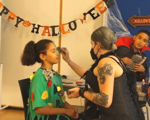 Kind in LernLEO wird von Betreuerin zu Halloween geschmückt, dahinter weiteres Kind in Dracula Kostüm, an Wand Happy Halloween Girlande