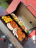 Ein Grill mit verschiedenen Grillwaren: mariniertes Fleisch, Würstchen, Gemüsespieße und Maiskolben. Der Grill ist geöffnet und zeigt eine bunte Auswahl an gegrilltem Essen. Im Hintergrund ist eine grüne Rasenfläche sichtbar.