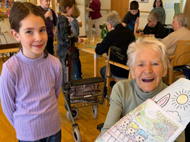 Ein junges Mädchen steht neben einer älteren Frau im Rollstuhl. Die Frau hält stolz ein bunt ausgemaltes Bild mit zwei Kindern und einer Landschaft. Im Hintergrund sind weitere Personen und Tische sichtbar, die auf eine kreative Aktivität hindeuten.