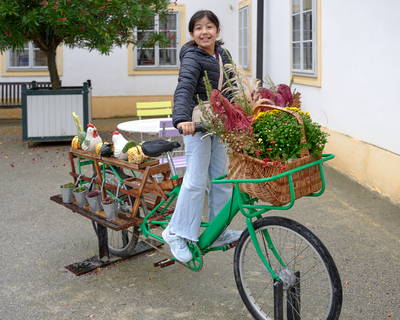 ein junges Mädchen sitzt auf einem umgebauten grünen Fahrrad auf dem Blumentöpfe über dem Hinterrad stehen, Generationenausflug