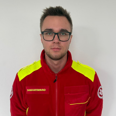 Ein junger Mann in einem roten, mit gelben Akzenten versehenen Feuerwehranzug steht vor einem neutralen Hintergrund. Er trägt eine Brille und hat einen ernsten Gesichtsausdruck. Auf seiner Uniform steht 