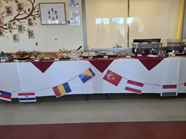 Ein reichhaltiges Buffet auf einem Tisch mit einer weißen Tischdecke und roten Details. Verschiedene Speisen sind angerichtet, umgeben von Flaggen anderer Länder, darunter die Türkei, Deutschland und Kroatien, die an einer Girlande befestigt sind.