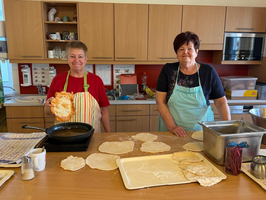 Zwei Frauen stehen in einer Küche, umgeben von Teigplatten und einer Pfanne. Eine Frau hält ein großes, gebackenes Fladenbrot in der Hand. An der Arbeitsfläche sind verschiedene Teigwaren und Küchengeräte sichtbar.