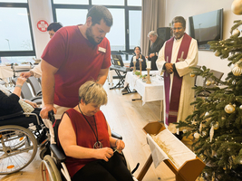 Weihnachtsfeier mit Menschen in Rollstühlen und einem Priester vor einem Christbaum.