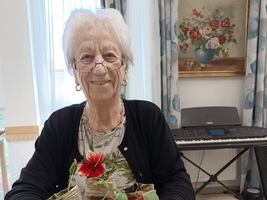 Eine ältere Frau lächelt, während sie eine große Schüssel mit Pflanzen, darunter frische Kräuter und eine rote Blume, in den Händen hält. Im Hintergrund sind ein Klavier und ein Blumenbild an der Wand zu sehen.