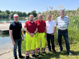 Fünf Männer stehen am Ufer eines Gewässers. Zwei tragen rote Arbeitskleidung mit gelben Hosen, während die anderen in Freizeitkleidung gekleidet sind. Im Hintergrund sind Bäume und Schilfrohr sichtbar. Es ist ein sonniger Tag.
