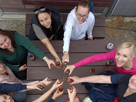 Eine Gruppe von zehn Personen steht um einen Tisch und stößt mit Getränken an. Die Menschen lächeln und haben diverse Kleidungsstücke an. Der Tisch ist mit Snacks und Getränken dekoriert.