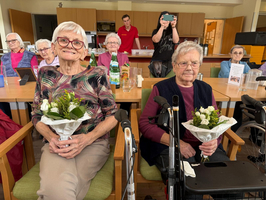 Zwei ältere Frauen sitzen in einem Gemeinschaftsraum und halten Blumensträuße in den Händen. Im Hintergrund sind weitere Seniorinnen zu sehen, die ebenfalls in der Gruppe sitzen. Ein junger Mann filmt die Szene mit einem Smartphone.