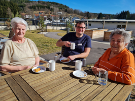 Drei Personen genießen Kaffee und Kuchen im Freien an einem Tisch.