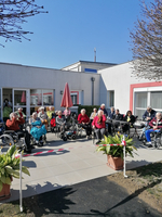 Ältere Menschen sitzen in einem Garten vor einem Gebäude, einige im Rollstuhl.