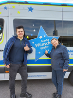 Zwei Personen neben einem Fahrzeug mit dem Schriftzug „Macht Wünsche wahr“.