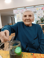 Eine ältere Frau in einem blauen Pullover sitzt an einem Tisch. Sie lächelt und hält einen Löffel in einer großen Glasflasche, die eine grüne Flüssigkeit enthält. Im Hintergrund sind Zeichnungen an der Wand sichtbar.