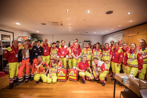 Eine Gruppe von Rettungskräften steht gemeinsam in einem Raum. Alle tragen auffällige rote und gelbe Uniformen. Einige Personen posieren für das Foto, während andere lächeln. Im Hintergrund sind Möbel und verschiedene Gegenstände sichtbar.