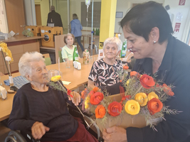Eine Frau überreicht einer Seniorin einen bunten Blumenstrauß in einem Gemeinschaftsraum.