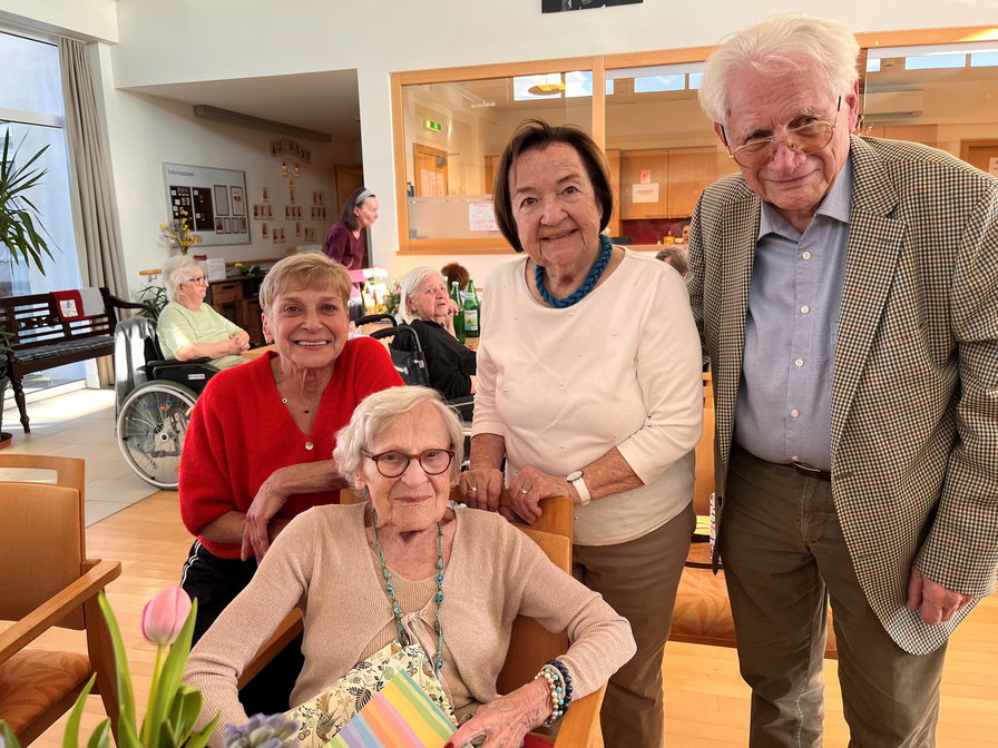 Eine Gruppe von vier älteren Menschen in einem Wohnheim. Drei stehen und lächeln, während eine Frau im Sessel sitzt. Auf dem Tisch sind Blumen und ein Notizbuch sichtbar. Im Hintergrund sind weitere Bewohner zu sehen.