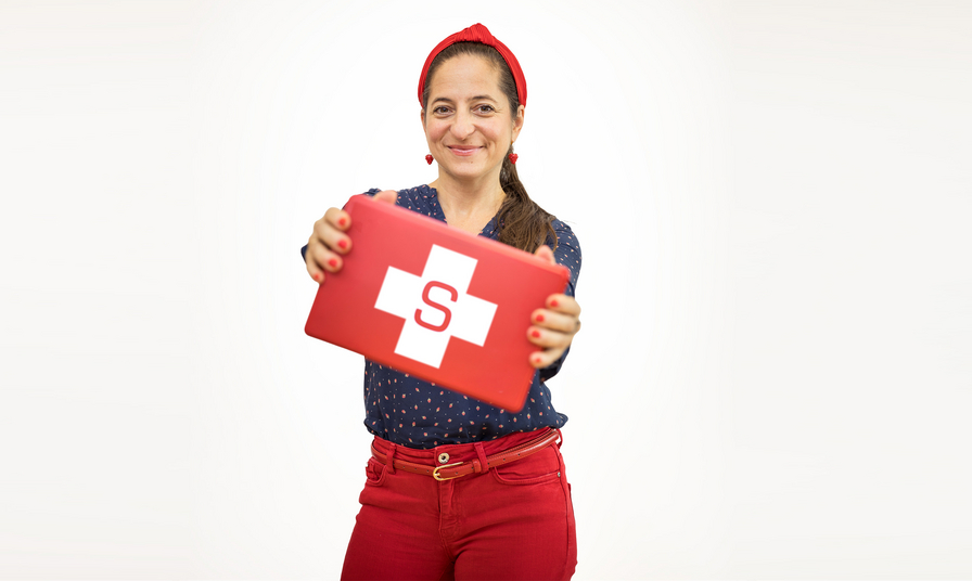 Eine lächelnde Frau mit einem roten Kopftuch und einem gepunkteten Oberteil hält eine rote Erste-Hilfe-Box mit einem weißen Kreuz und einem 