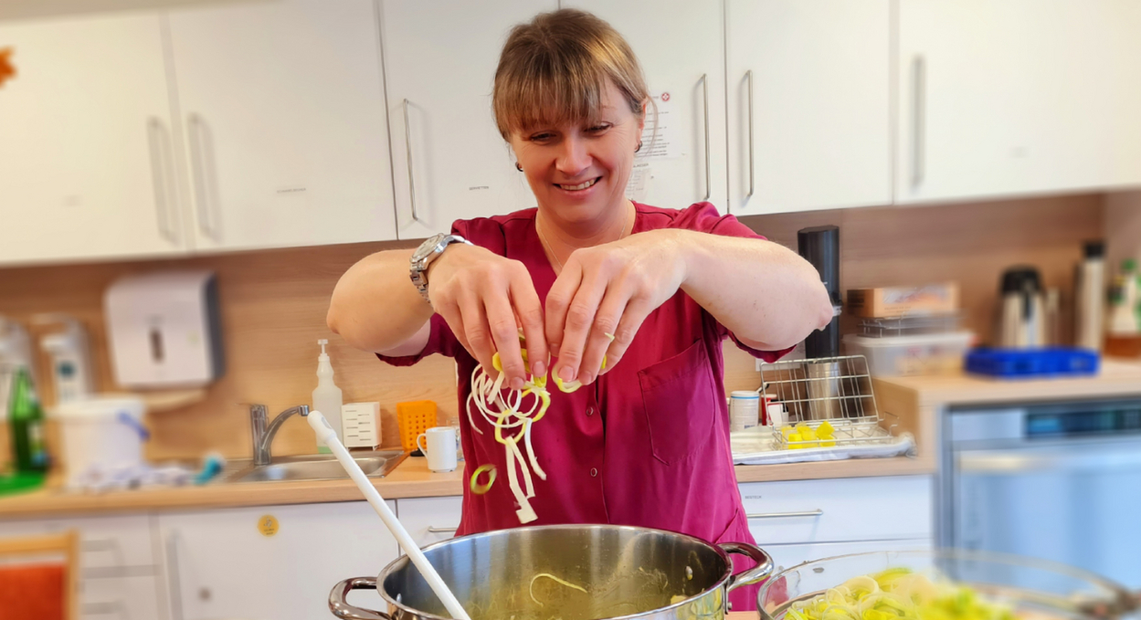 Eine Frau in einem roten Oberteil steht in einer Küche und fügt frisch geschnittene Lauchringe in einen Kochtopf. Neben dem Topf steht ein weiterer Topf mit Wasser und eine Schüssel mit geschnittenem Gemüse.