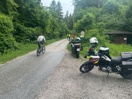 Fahrradfahrer und Motorräder auf einem schmalen Weg umgeben von Bäumen.