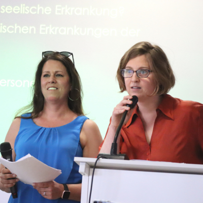 Zwei Frauen stehen vor einer Leinwand. Die Frau auf der linken Seite hält ein Mikrofon und Zetteln in der Hand, die Frau auf der rechten Seite steht vor einem Pult und spricht in ein Mikrofon.