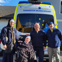 Vier Personen, darunter eine Schwester, stehen neben einem Rollstuhl und einem Fahrzeug.