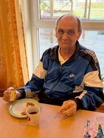 Ein älterer Mann sitzt an einem Tisch und lächelt. Er trägt eine blaue Sportjacke und hält ein Stück Kuchen in der Hand. Vor ihm steht ein Teller mit Kuchenstücken und eine Tasse Tee. Im Hintergrund sind Fenster und eine freundliche Atmosphäre sichtbar.