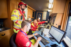 Vier Rettungskräfte arbeiten in einem mobilen Einsatzraum. Zwei sitzen an Computern und tippen, während zwei andere stehen und auf Bildschirme zeigen. Alle tragen rote und gelbe Arbeitskleidung. Der Raum ist mit technischen Geräten ausgestattet und hat holzpaneelierte Wände.