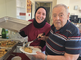 Eine lächelnde Frau in einem roten Oberteil serviert Essen aus einem Tablett, während ein älterer Mann in gestreiftem Hemd einen Teller mit einer Portion aufnimmt. Im Hintergrund sind weitere Gerichte und Getränke sichtbar.