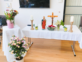 Ein festlich dekorierter Tisch mit weißen Tischdecken. Auf dem Tisch stehen eine blühende Pflanze, eine Statue, Kerzen und eine Schale mit Blumen. Im Hintergrund sind farbige Blumen in einem Topf und ein Fernseher zu sehen.