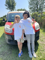 Zwei Kinder in weißen T-Shirts posieren lächelnd vor einem Fahrzeug.