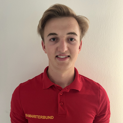 Ein junger Mann mit blonden Haaren trägt ein rotes Polo-Shirt mit der Aufschrift „Samariterbund“. Er steht vor einer weißen Wand und lächelt.