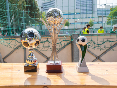 drei Pokale mit unterschiedlichem Aussehen, alle im Zusammenhang mit Fußball, stehen auf Biertisch, dahinter Netz eines Fußballfelds, im Hintergrund Spieler erkennbar, Bäume, Gebäude