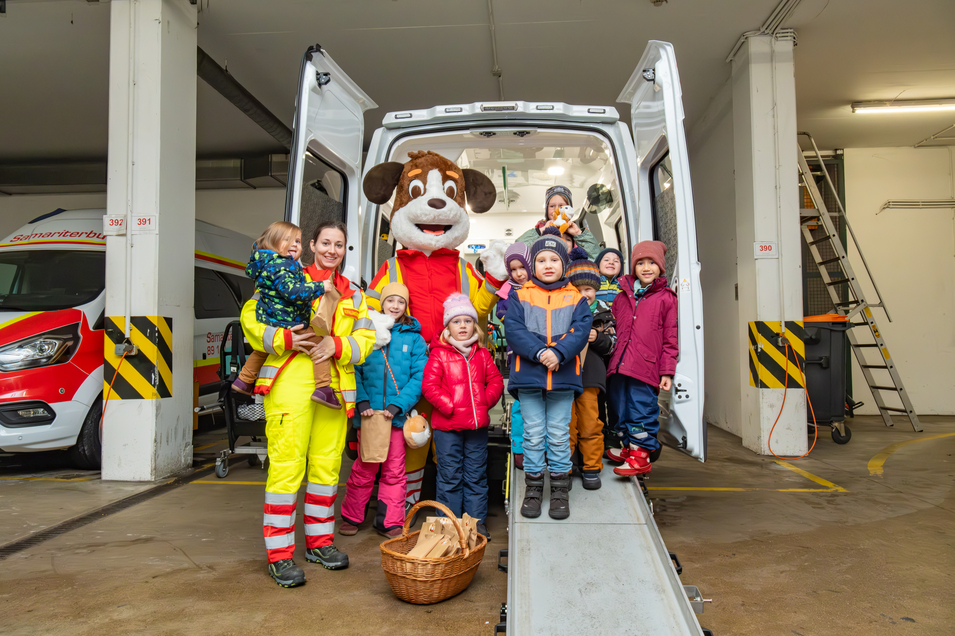 Kindergruppe mit Maskotchen Sam und einer Sanitäterin vor einem Rettungsfahrzeug vom Samariterbund Wien