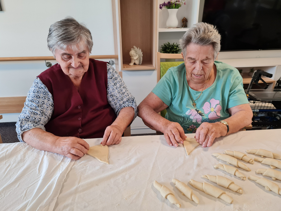 Zwei ältere Frauen sitzen an einem Tisch und rollen Teig zu kleinen Formen. Vor ihnen liegen bereits einige vorbereitete Teigstücke. Beide konzentrieren sich auf ihre Arbeit und arbeiten in einer freundlichen, gemeinschaftlichen Umgebung.