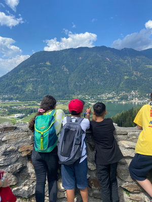 Drei Kinder stehen mit dem Rücken zur Kamera vor einer Steinwand. Im Hintergrund sieht man einen großen Berg und einen See.
