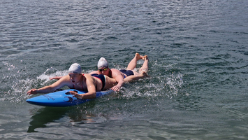 Zwei Schwimmer paddeln auf einem Surfbrett im Wasser.