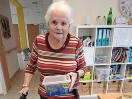 Eine ältere Frau mit Rollator hält eine Box in der Hand.