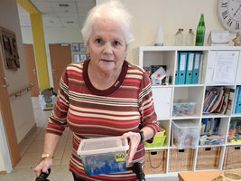 Eine ältere Frau mit Rollator hält eine Box in der Hand.