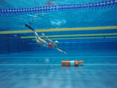 Eine Schwimmerin taucht unter Wasser in einem Schwimmbecken. Sie trägt eine rote Badeanzug und eine Schwimmkappe. Neben ihr befindet sich eine orangefarbene Boje. Die Unterwasseransicht zeigt die Klarheit des Wassers und die Schwimmbeckenmarkierungen im Hintergrund.
