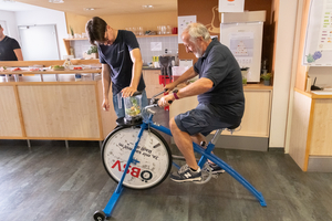 Person sitzt auf einem umgebauten Fahrradgestellt und betreibt durch das Treten einen Smoothie-Mixer, andere Person steht vor Rad und hält Mixer fest